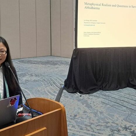 iamge of Lu Huang standing at a podium presenting and smiling at the camera with her paper displayed on an overhead projector behind her