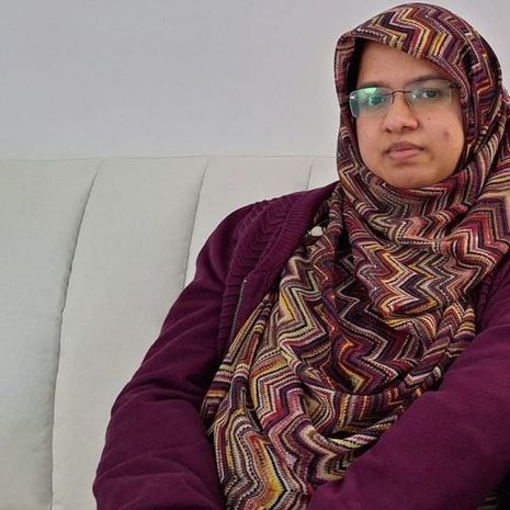 image of Hamida sitting on a couch wearing glasses and a purple garb and geometric pattern hijab