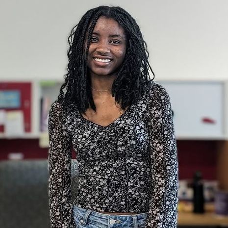 image of Imanee standing in front of desks in the Temple newsroom smiling and wearing a black and white print blouse and jeans