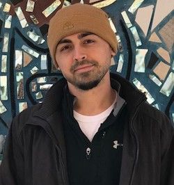image of Jake standing against a mosaic wasll weaing a brown knit cap and black jacket