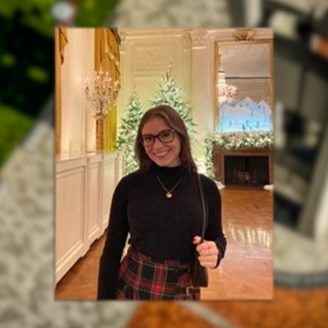 image of Kira standing with Christmas trees in the background wearing glasses, a black turtleneck and a red plaid skirt and holding her purse