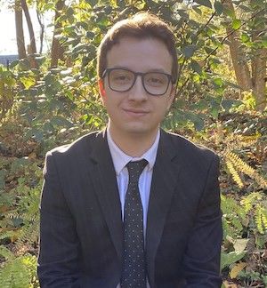 image of Davisd wearing glasses and a black suit and blue tie sitting witth the forest behind him
