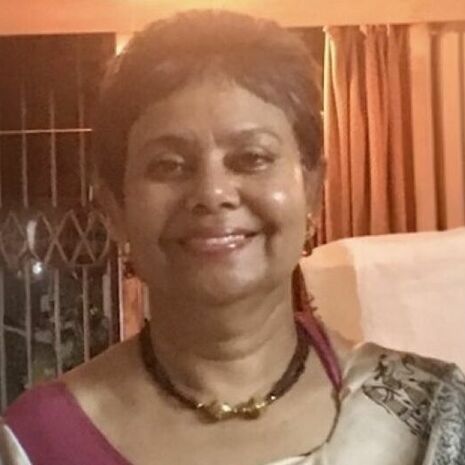 image of Srimati Mukherjee standing and smiling at the camera wearing a gold and brown necklace and light tan and maroon saree