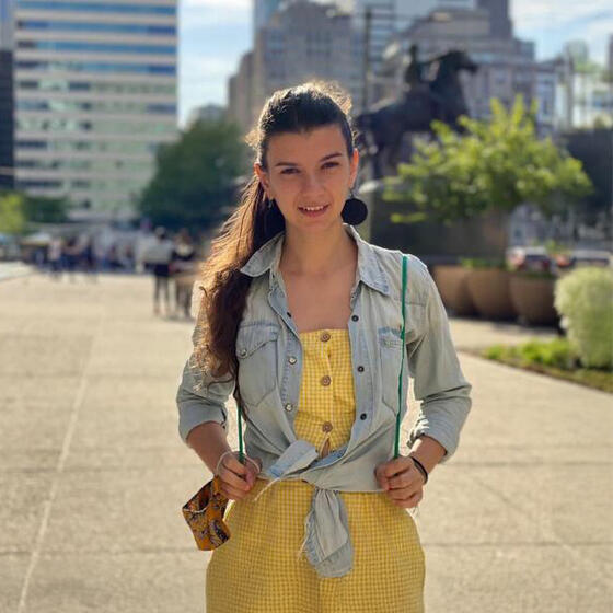 Angela Mejia in a yellow jump suit and jean jacker standing outside staring into the camera