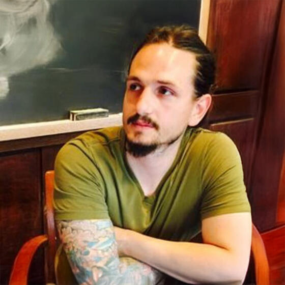 Alex Melonas in a classroom with his arms crossed staring off