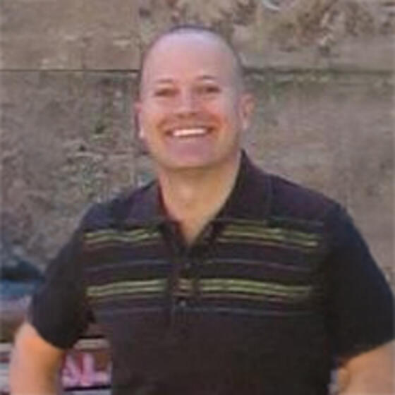 Michael Bognanno wearing a black shirt standing in front of a wall smiling and starting into the camera