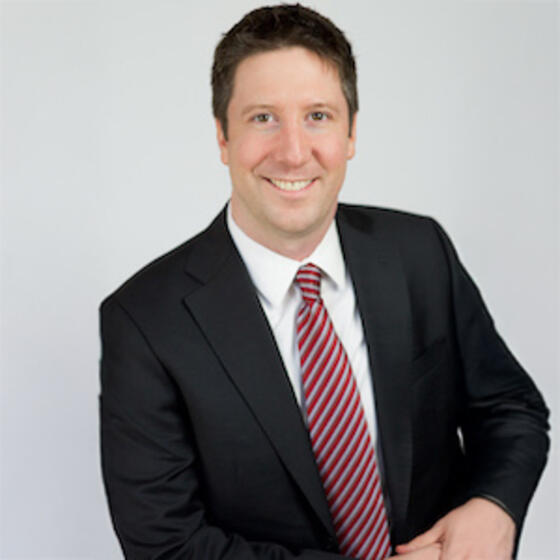 Joshua Mask wearing a black suit and red tie staring into the camera and smiling