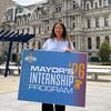  image of Luana standing oil front of City Hall golding a Mayors Internship sign and smiling at the camera