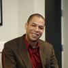 Kareem Johnson in a brown suit coat and red shirt smiling and staring into the camera