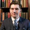 Alexander Falbo-Wild in a black suit with a blue and black stripped tie in front of a book shelf
