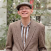 Austin Fuller outside in a brown suit coat and hat on smiling into the camera