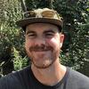 image of Brookes standing in the woods wearing a camoflauge hat and black tee shirt smiling at the camera
