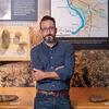 image of Greg standing with his arms crossed against a table of artifacts wearing denim and a blue shirt and glasses looking at the camera