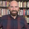 image of Julien wearing a plaid shirt standing in front of a bookcase smilng at the camera