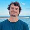 Kyle McDonald standind in front of an ocean in a blue shirt smiling into the camera