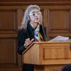 Laura Levitt speaking at a podium with a black blazer and colorful scarf talking to the crowd