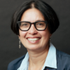 image of Priya wearing glasses and a grey suit smiling at the camera