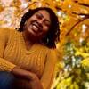 image of Yesinia wearing a gold sweater smiling at the camera in front of a tree with orange and yellow colored leaves