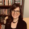 image of Courtney wearing glasses and a black top sitting in from of a bookcase smiling at the camera