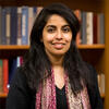 Aunshul Rege standing in front of a bookshelf wearing a black shirt with a colorful scarf looking into the camera