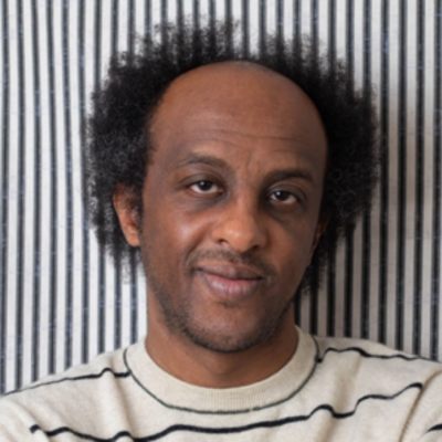 image of Dinaw standing against a black and white striped wall weaing a black and white striped shirt half smiling at the camera