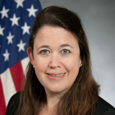 image of Heather wearing a black suit standing in front of an American flag, smiling at the camera