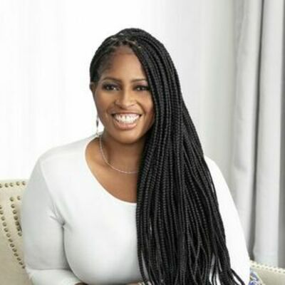 image of Mecca sitting in a tan chair wearing a white top with long braids covering half her shoulder smiling at the camera