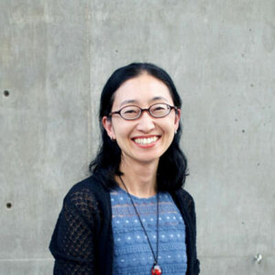 imgae of Sawako wearing a blue top and black cardigan, wearing glasses and smiling at the camera