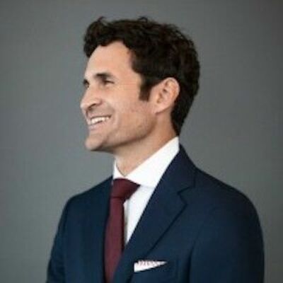 image of Gus smiling and looking off into the distance, wearing a blue suit and maroon tie