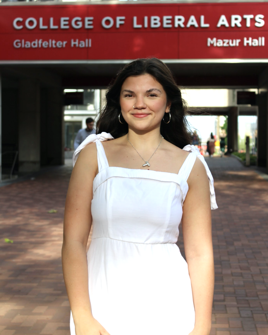 image of Haleigh standing in a white dress smiling in front of the red and white College of Liberal Arts sign