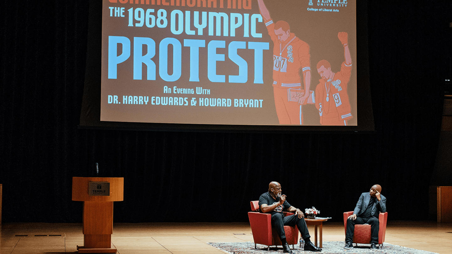 Dr. Harry Edwards and Howard Bryant sit on a stage in front of a screen that reads 1968 olympic protest