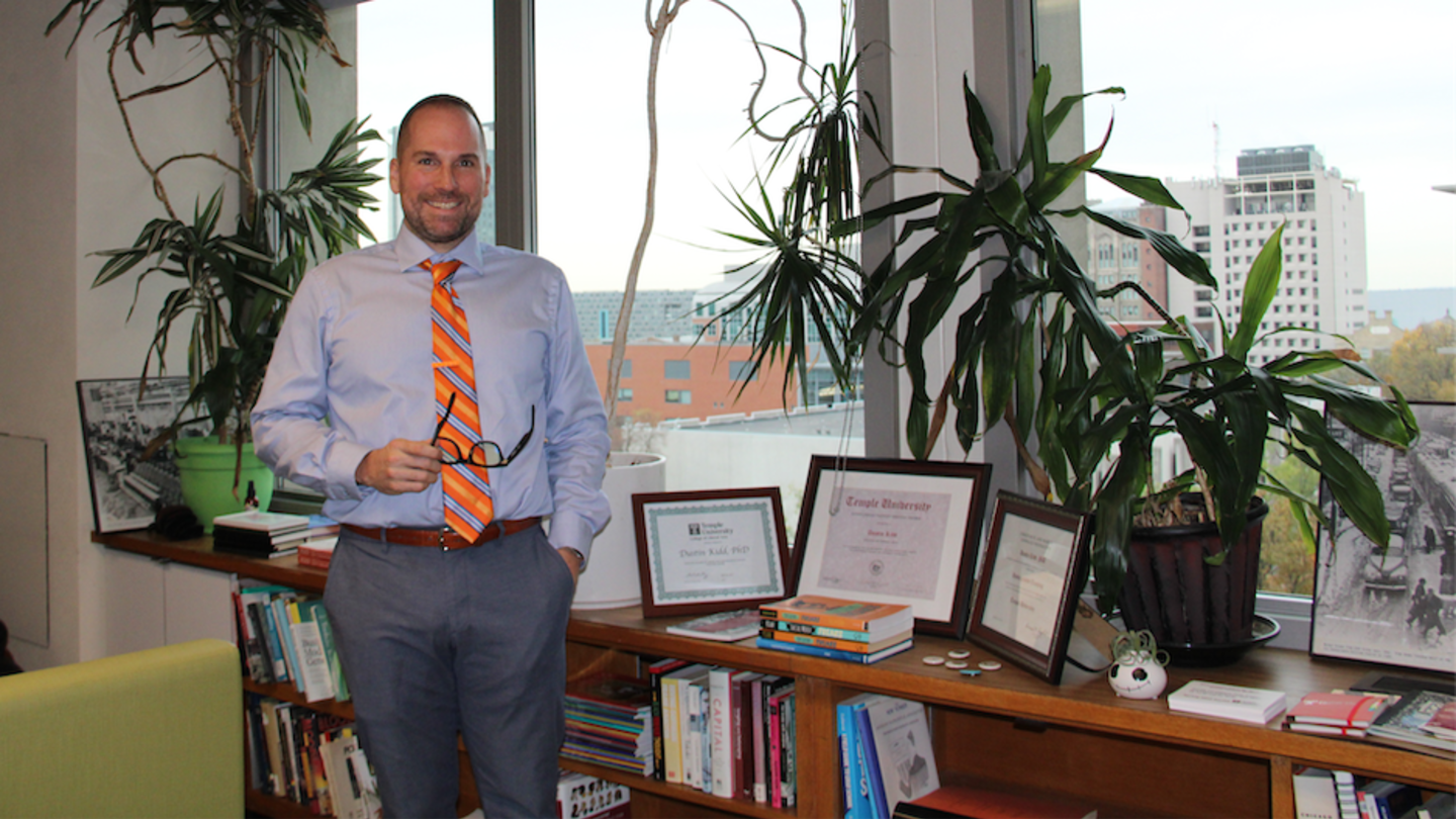 image of Dustin Kidd standing in his office in a blue shirt, grey pants and an orange tie