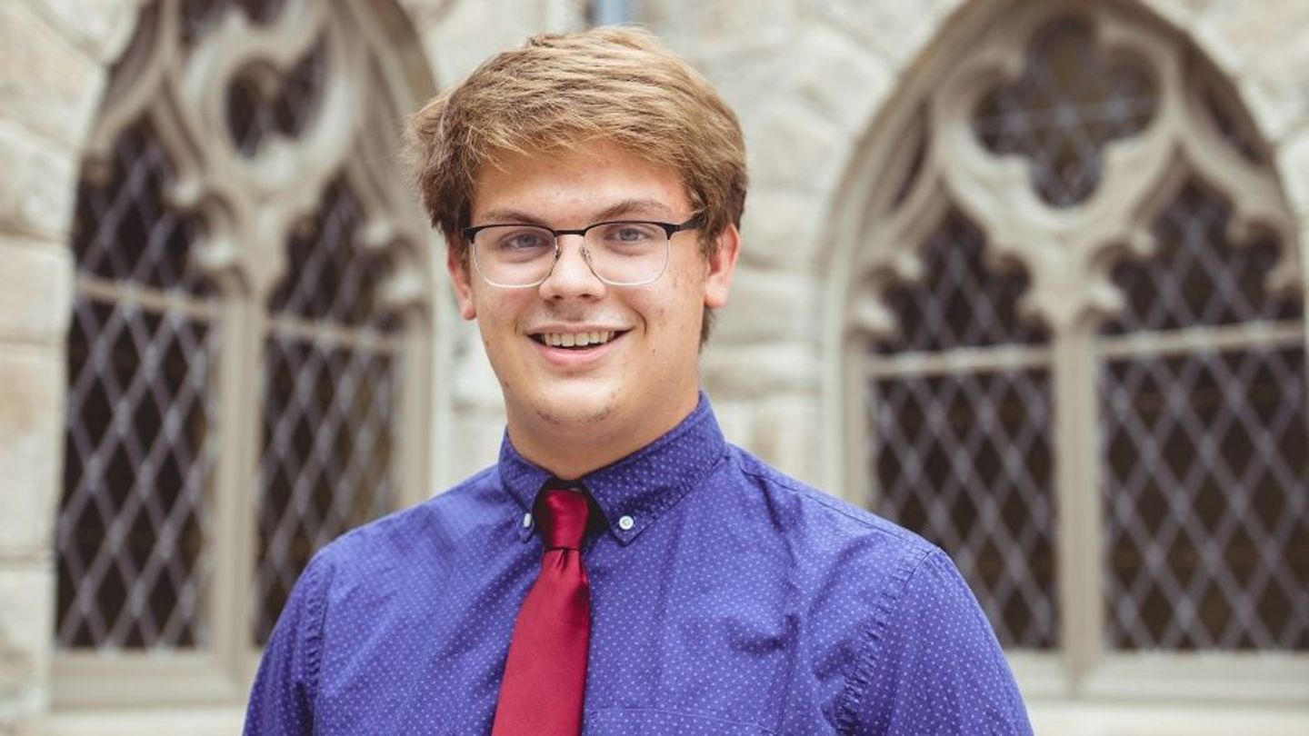 image of Sam Hall in a blue shirt and red tie standing in front of a cathedral