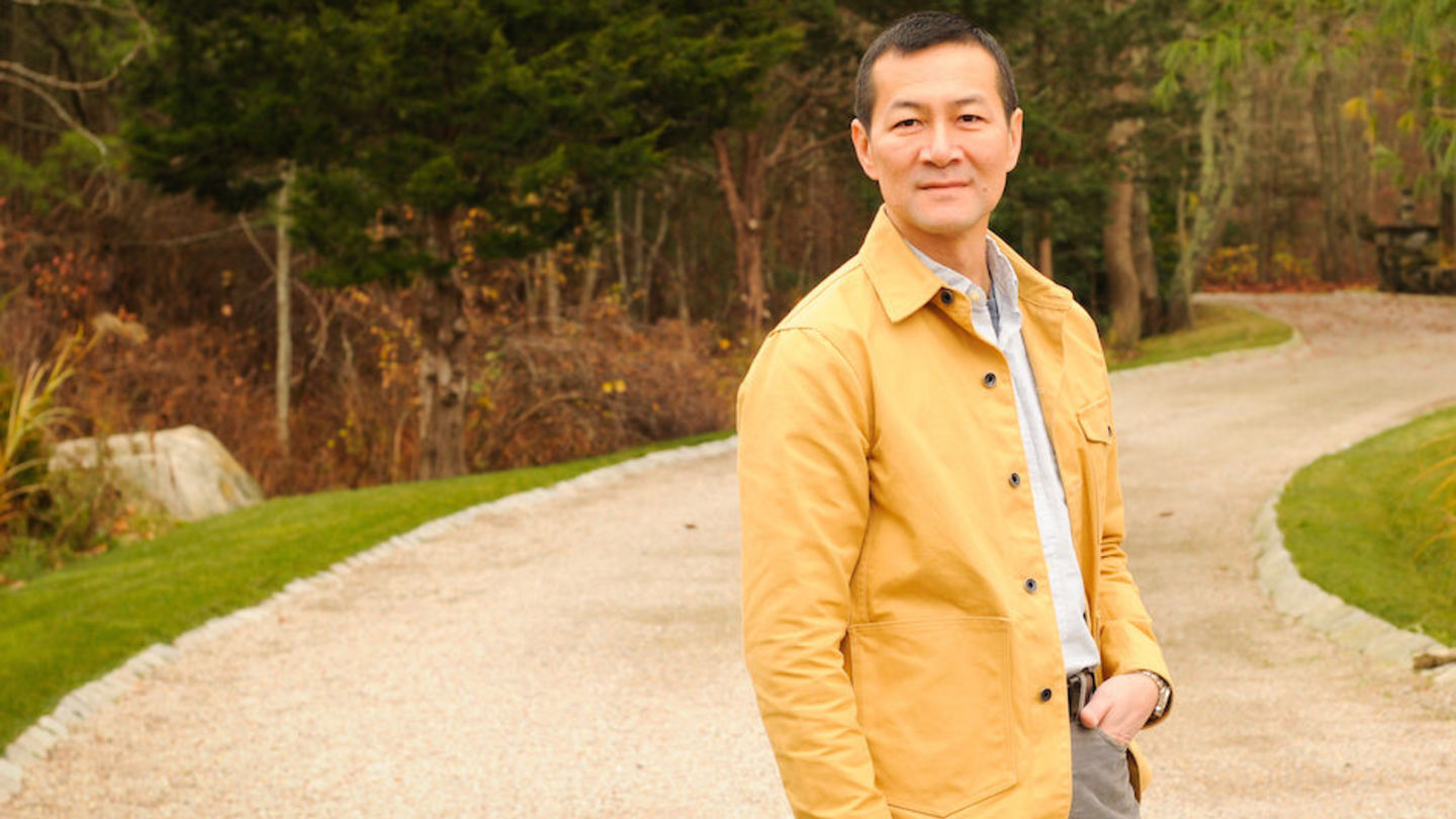 image of Don Lee wearing a yellow coat, white shirt, grey pants standing in a road with the forest in the background