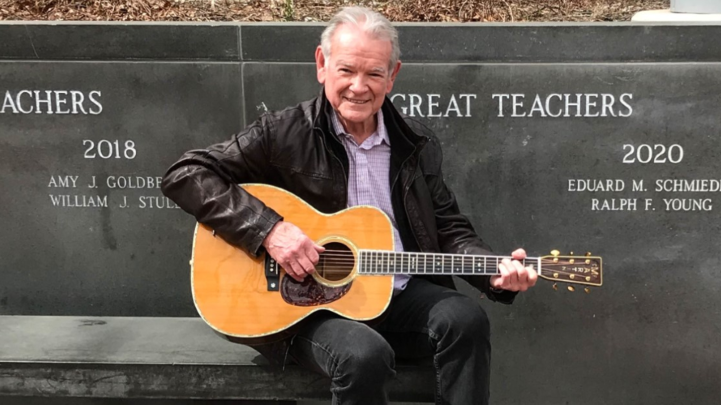 image of Ralph sitting in founders garden, playing his guitar in front of his name on the great teachers award wall