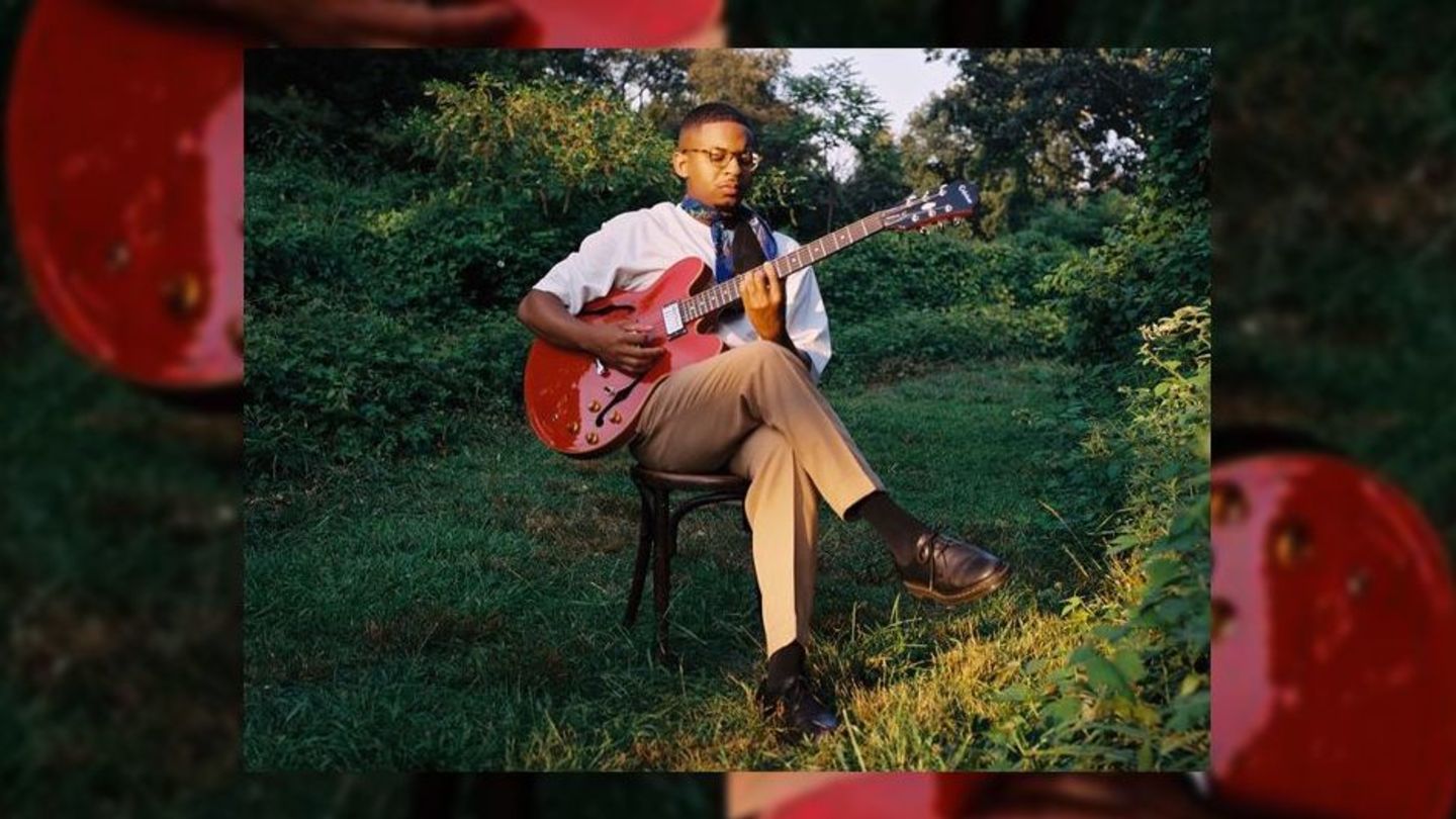 image of Stanley, sitting in a white tee shit and tan pants with his legs crossed playing a red guitar in a wooded area