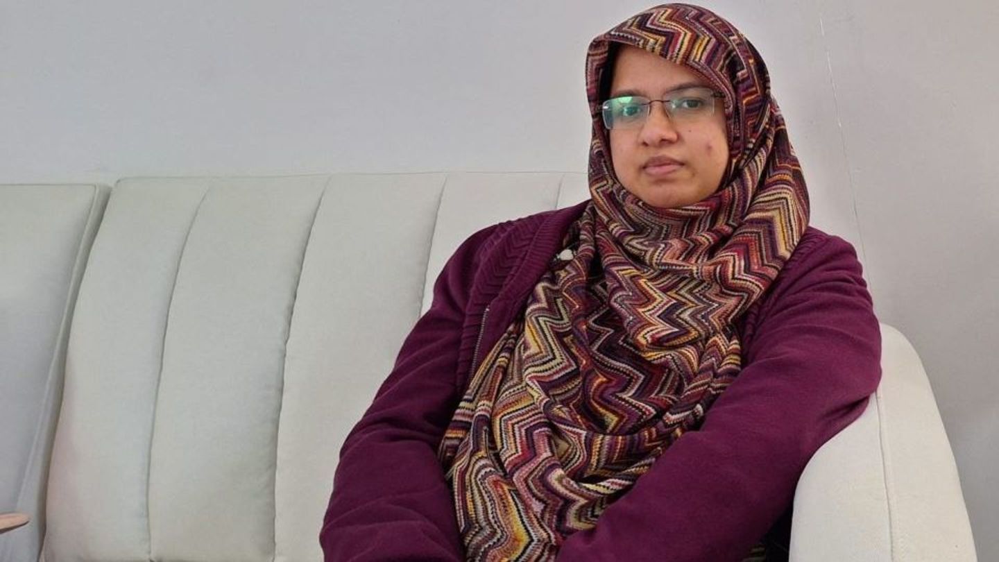 image of Hamida sitting on a couch wearing glasses and a purple garb and geometric pattern hijab