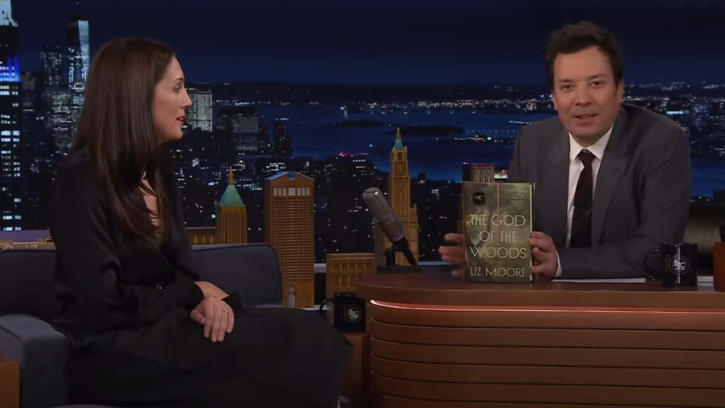 image of Liz Moore sitting in a blue dress looking at Jimmy Kimmel as he holds her book up towards the camera