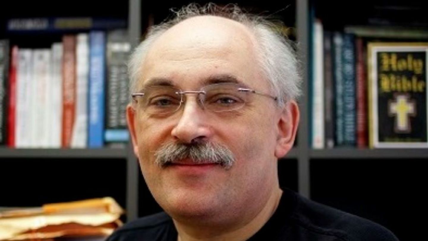 image of Mikhail sitting at his desk wearing a black t-shirt and glasses smiling at the camera