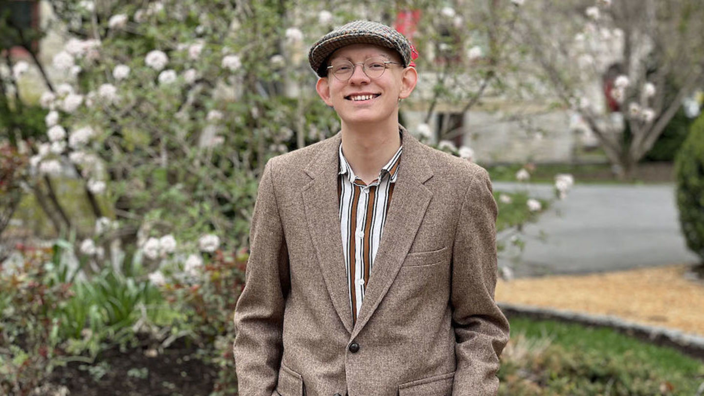 image of Austin standing in front of blooming flowers wearing glasses, a brown suit jacket, a hat and striped shirt