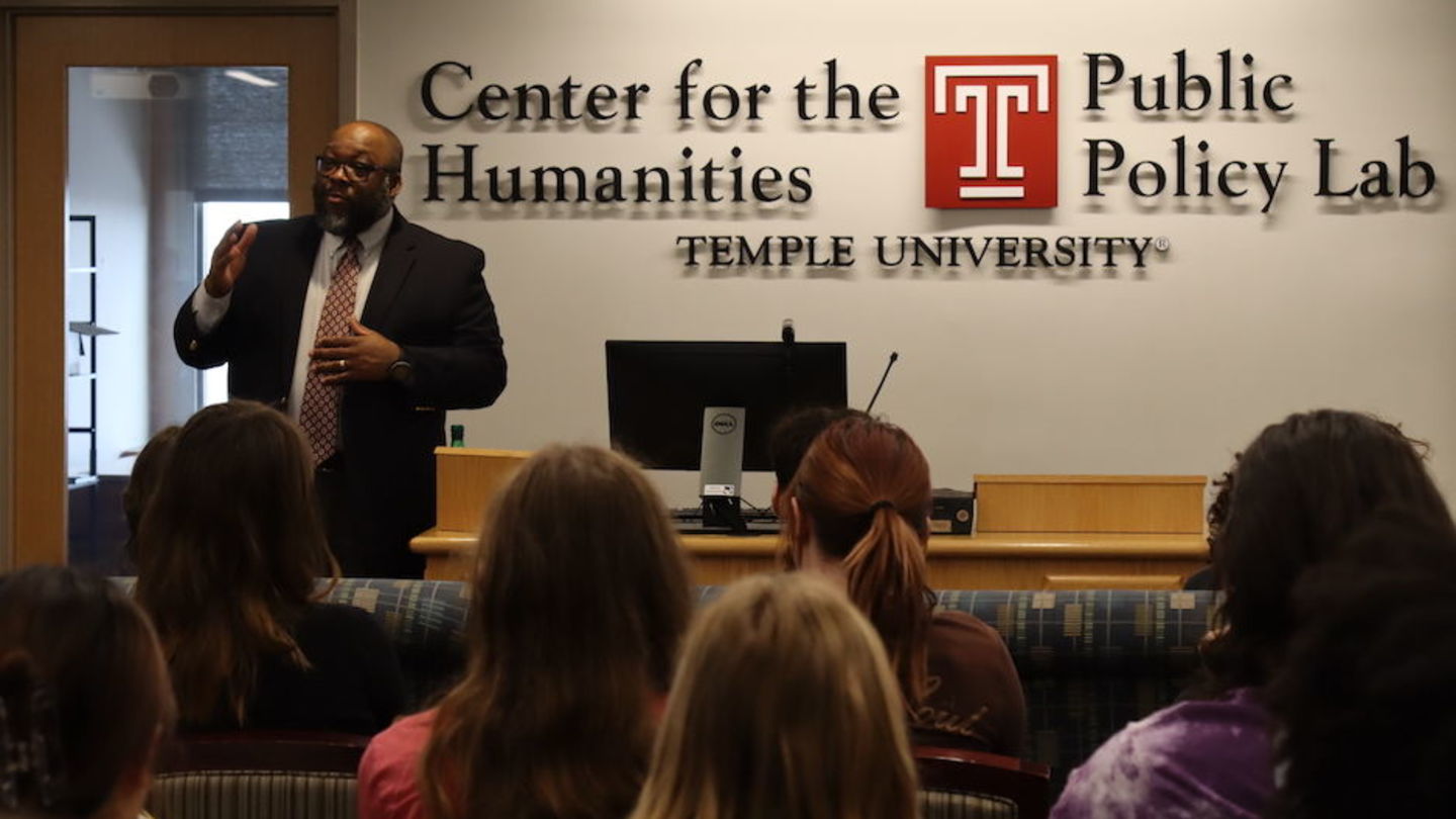 image of Art standing in front of students wearing glasses and a suit speaking to them