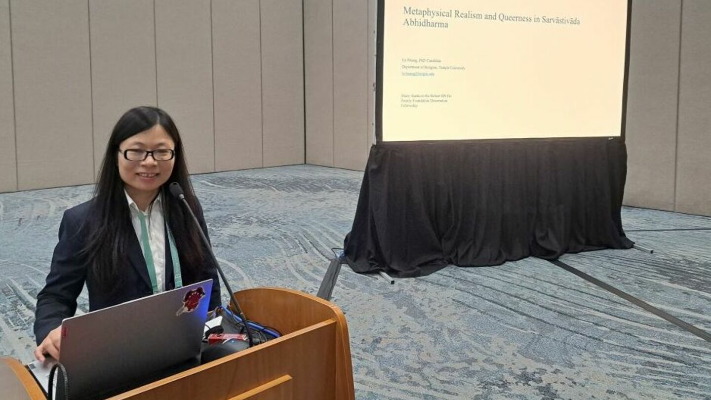 iamge of Lu Huang standing at a podium presenting and smiling at the camera with her paper displayed on an overhead projector behind her