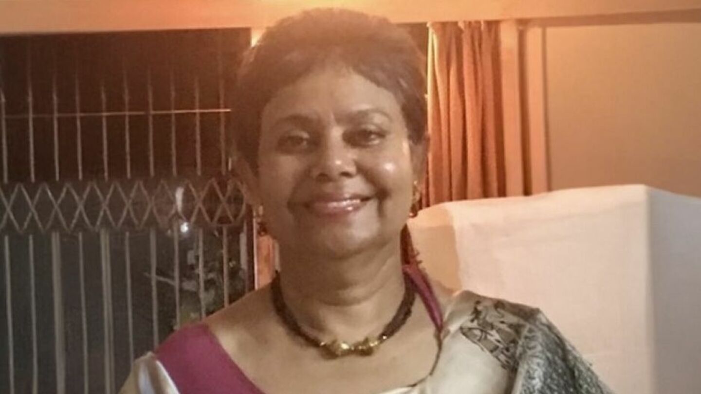 Professor Srimati Mukherjee image of Srimati Mukherjee standing and smiling at the camera wearing a gold and brown necklace and light tan and maroon saree